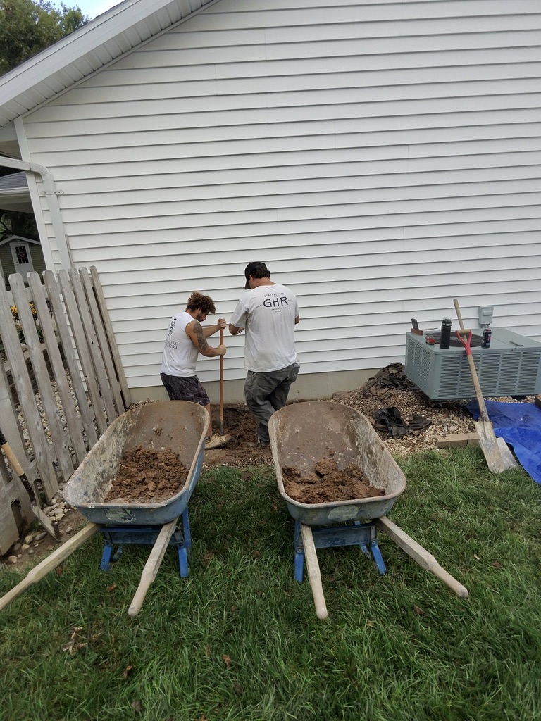 GHR Egress Windows crew hand-digging an egress window excavation beside a house in Waterloo, Illinois, with two wheelbarrows of dirt