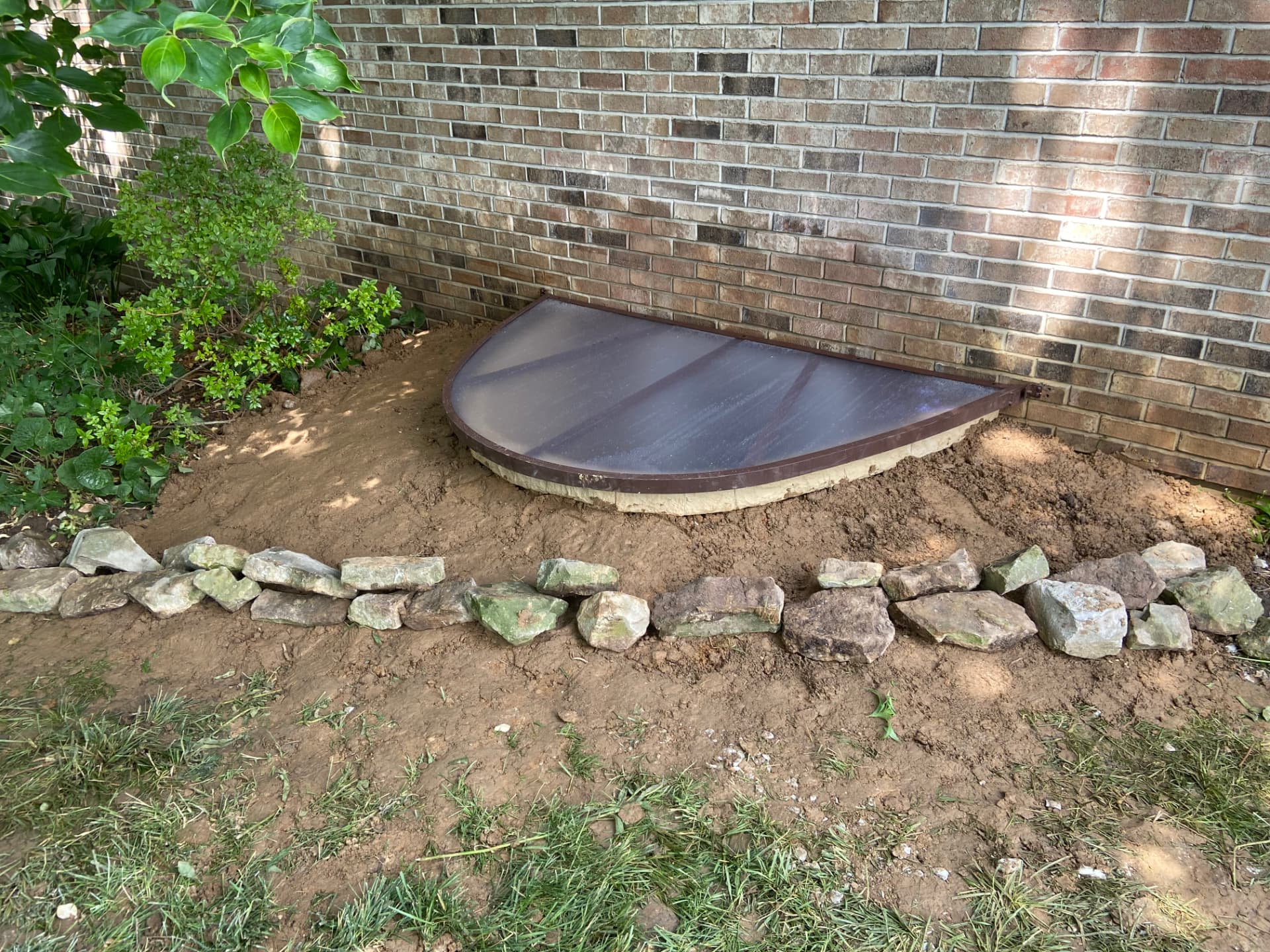 Finished egress window well with a brown framed clear cover against a brick foundation, installed by GHR Egress Windows.