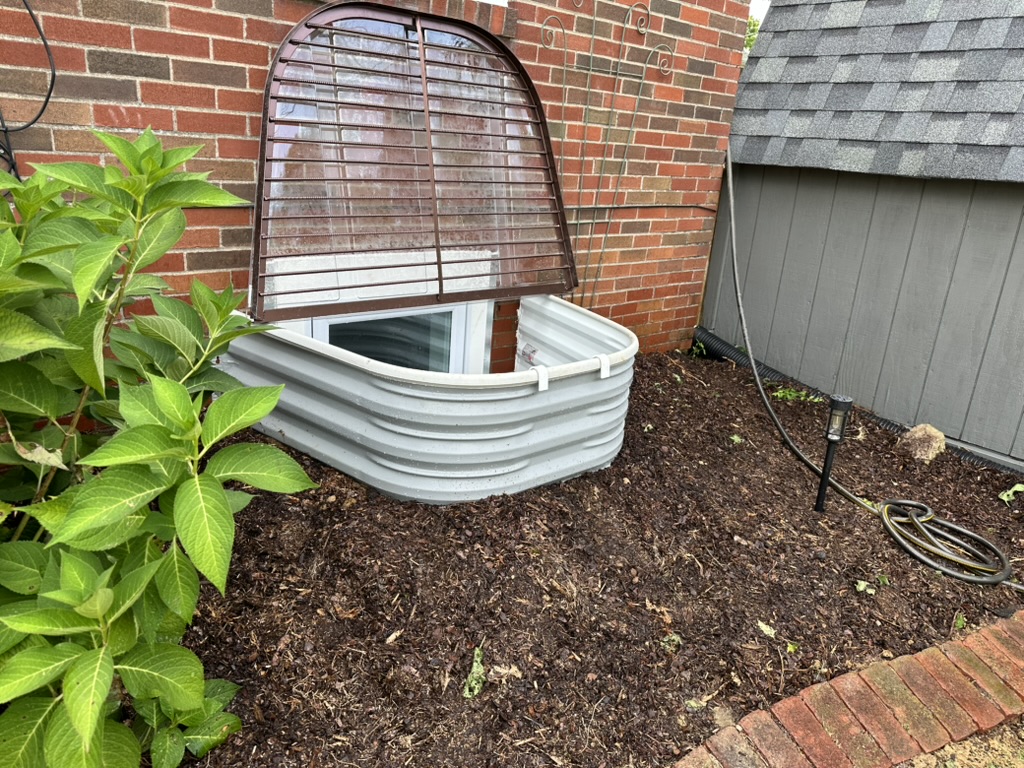 Exterior view of a white steel egress window well with grate and clear cover installed by hand in Edwardsville, Illinois, by GHR Egress Windows.